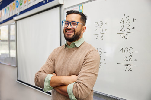 Happy teacher explaining mathematics at whiteboard in classroom. Portrait of smiling mathematics tutor explaining addition concept to children in classroom. Successful teacher standing in classroom.