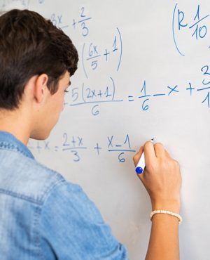 Back view of high school student solving math problem on whiteboard in classroom. Young man writing math solution on white board using marker. College guy solving math expression during lesson.