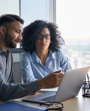 Indian male ceo executive manager mentor giving consultation on financial operations to female African American colleague intern using laptop sitting in modern office near panoramic window.