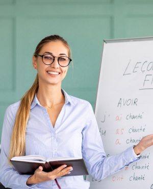 Happy French teacher explaining foreign language rules near blackboard indoors and smiling at camera. Confident young female tutor teaching linguistic online course