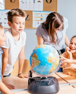 Happy cute enthusiastically pupils smiling around a globe in classroom with female teacher at school. Group of curious classmates at workplace studying globe in classroom.