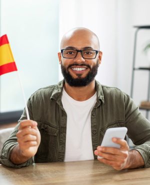 Excited male tutor sitting at table with flag of Spain and using smartphone, looking and smiling at camera, copy space. Positive latin man studying spanish language, emigration to Europe concept