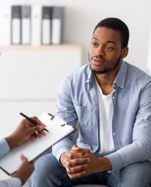 Unhappy young black man having session with professional psychologist at mental health clinic. Psychotherapist taking notes during conversation with depressed male patient
